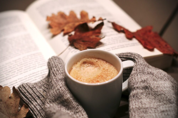 Book covered with leaves behind a sweater holding a cup of coffee